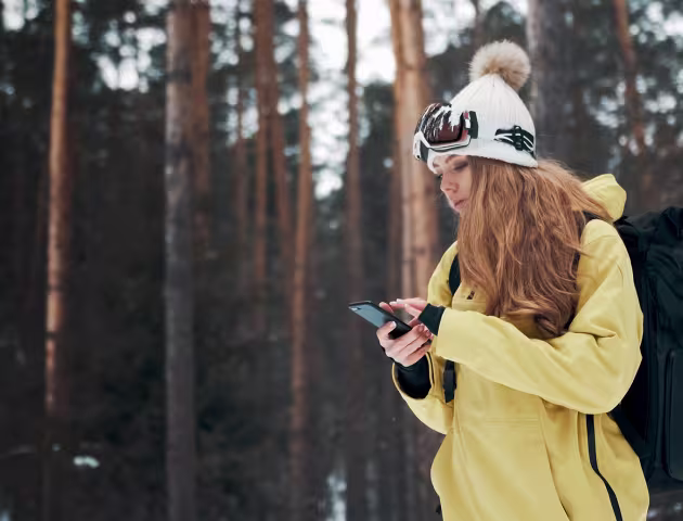 Dziewczyna w żółtej kurtce i czapce na tle lasu. Sprawdza coś w telefonie.