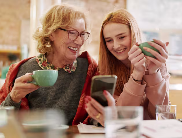 Babcia i wnuczka patrzą na telefon komórkowy, pijąc herbatę w kuchni