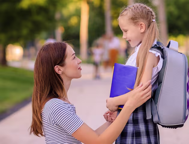 mama kuca i trzyma za ręce córkę z tornistrem, patrza na siebie z uśmiechami
