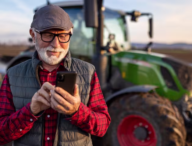 Pan z siwą brodą z komórką, w tle traktor
