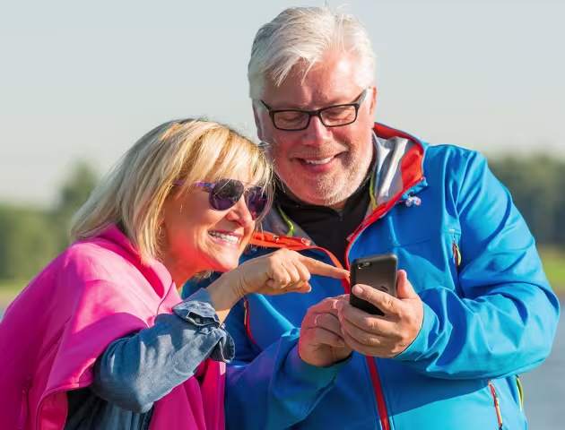 Para w średnim wieku w kurtkach nad wodą, pokazuja sobie coś na telefonie