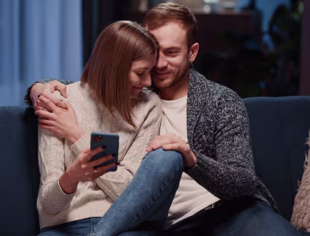 Przytulona para na kanapie, ona trzyma w ręku komórkę