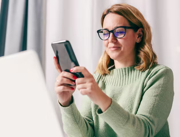 30latka z komórką, na którą patrzy, przed nią laptop