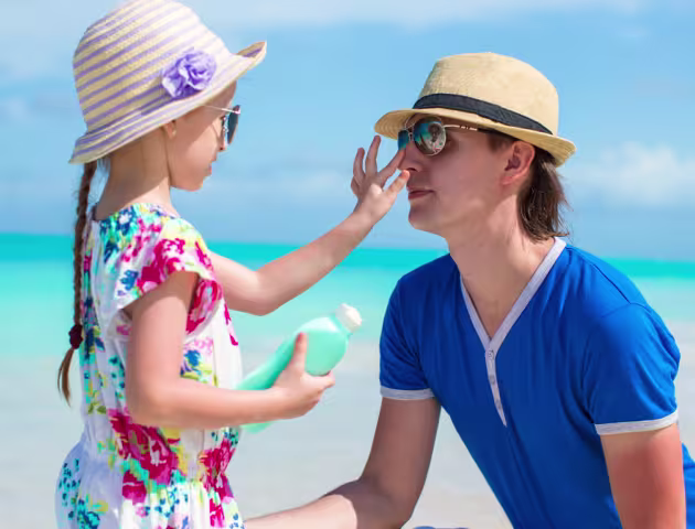 Ojciec i córka na plaży, oboje w słomkowych kapeluszach i okularach przeciwsłonecznych. Dziewczynka trzyma butelkę, drugą ręką dotyka nosa ojca.