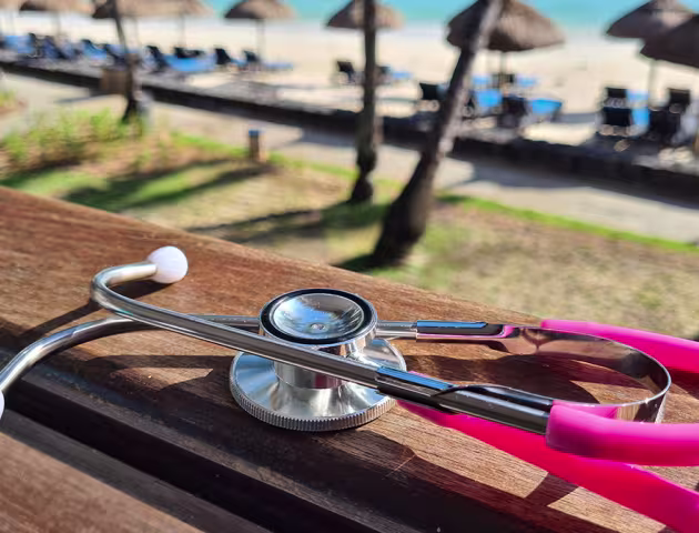 Różowy stetoskop leży na ławce w słońcu, w tle palmy i parasole na plaży