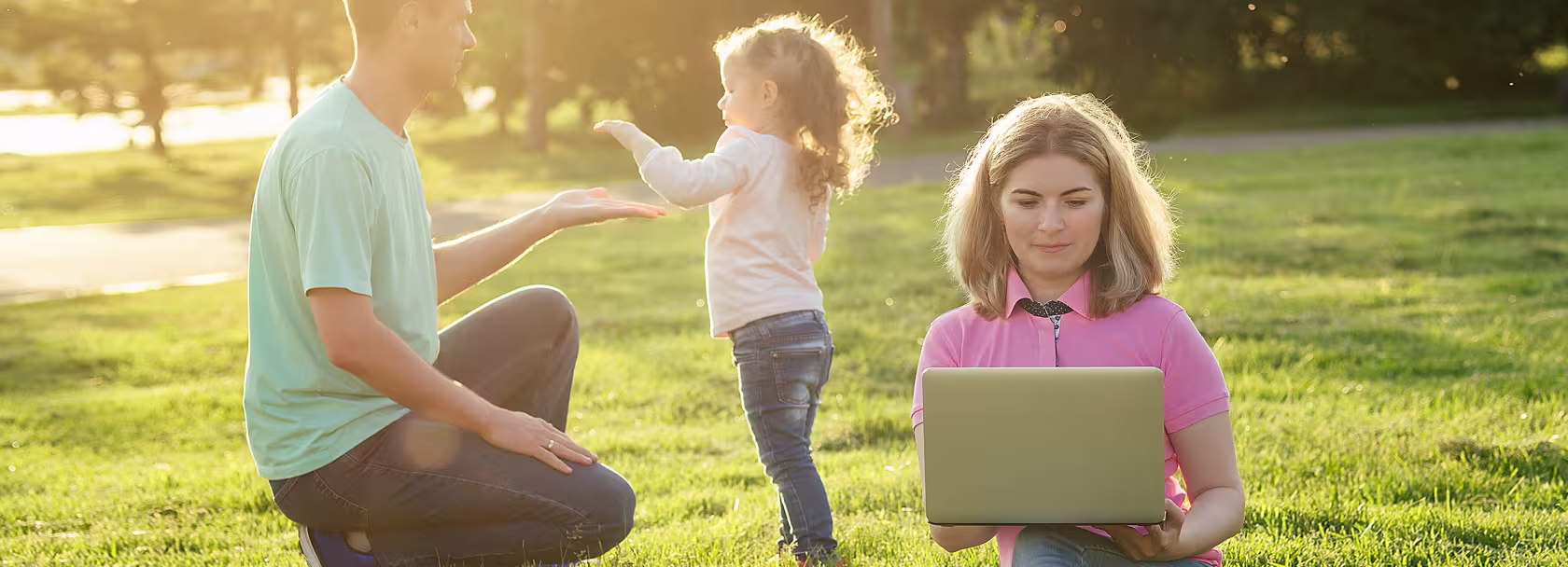 Ojciec bawi się z córeczką na łące, matka zagląda do laptopa