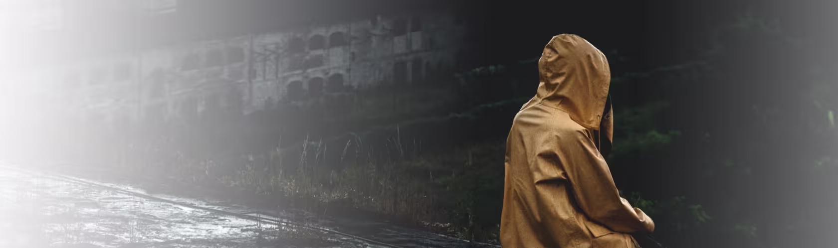Siedząca tyłem na zaniedbanym wyłomie budynku osoba w żółtym płaszczu przeciwdeszczowym, z kapturem na głowie