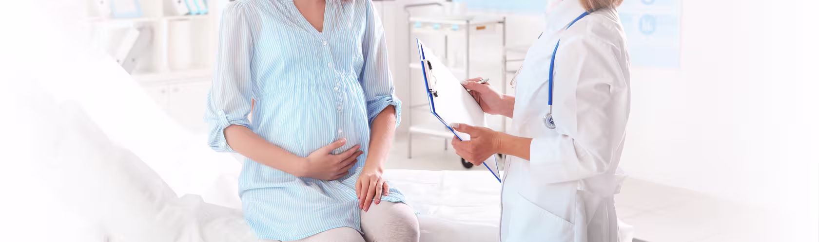 Młoda, uśmiechnięta kobieta w ciąży siedzi na kozetce i trzyma się za brzuch podczas rozmowy z lekarką.