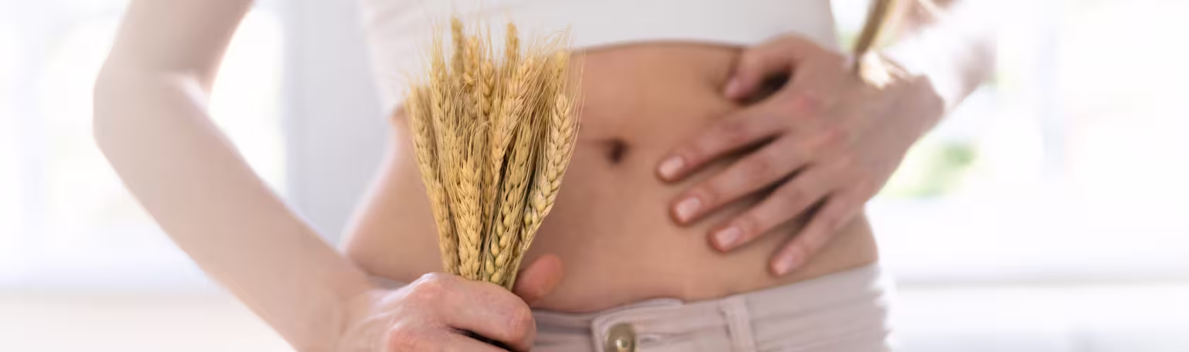 Zbliżenie na goły brzuch kobiety. W jednej ręce trzyma kłosy zbóż, a drugą dotyka brzucha.