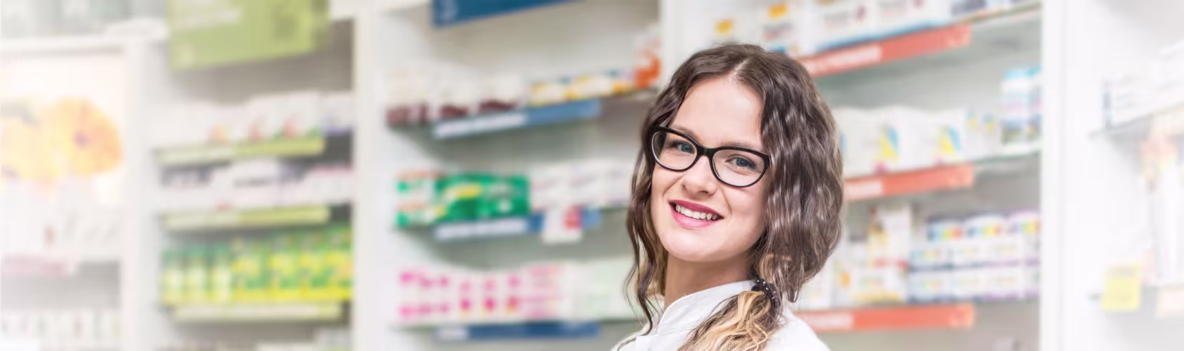 Uśmiechnięta farmaceutka w aptece. Młoda dziewczyna ma długie, falowane włosy upięte w kucyk na boku. Ma okulary.