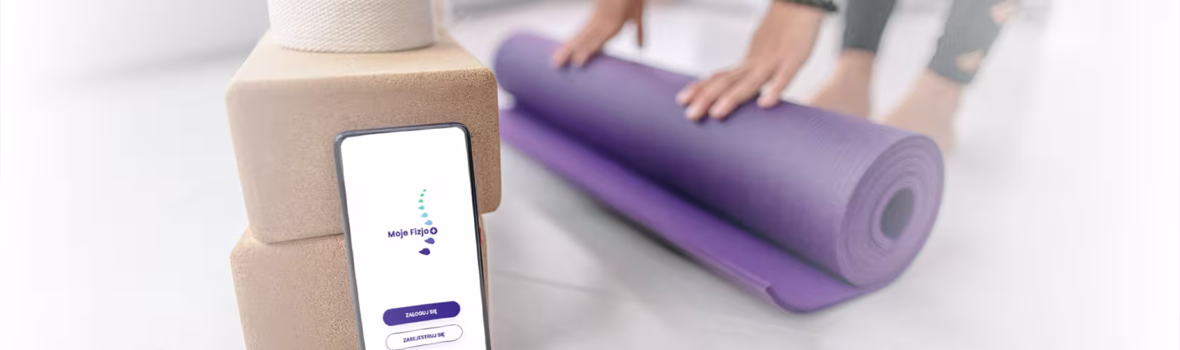 Smartfon z aplikacją Moje Fizjo na ekranie. W tle ręce rolujące matę do ćwiczeń.