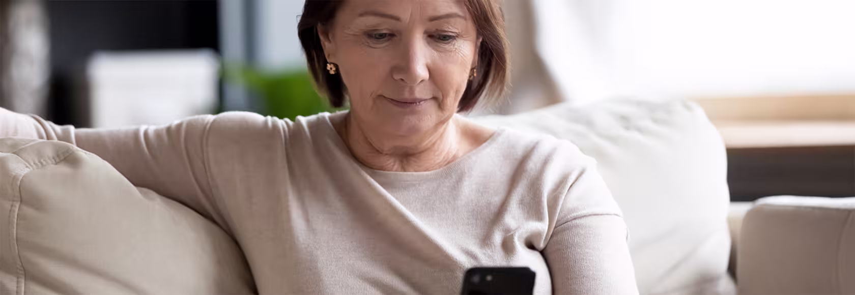 Pani w beżowym t-shircie patrzy na telefon, siedzi na kanapie
