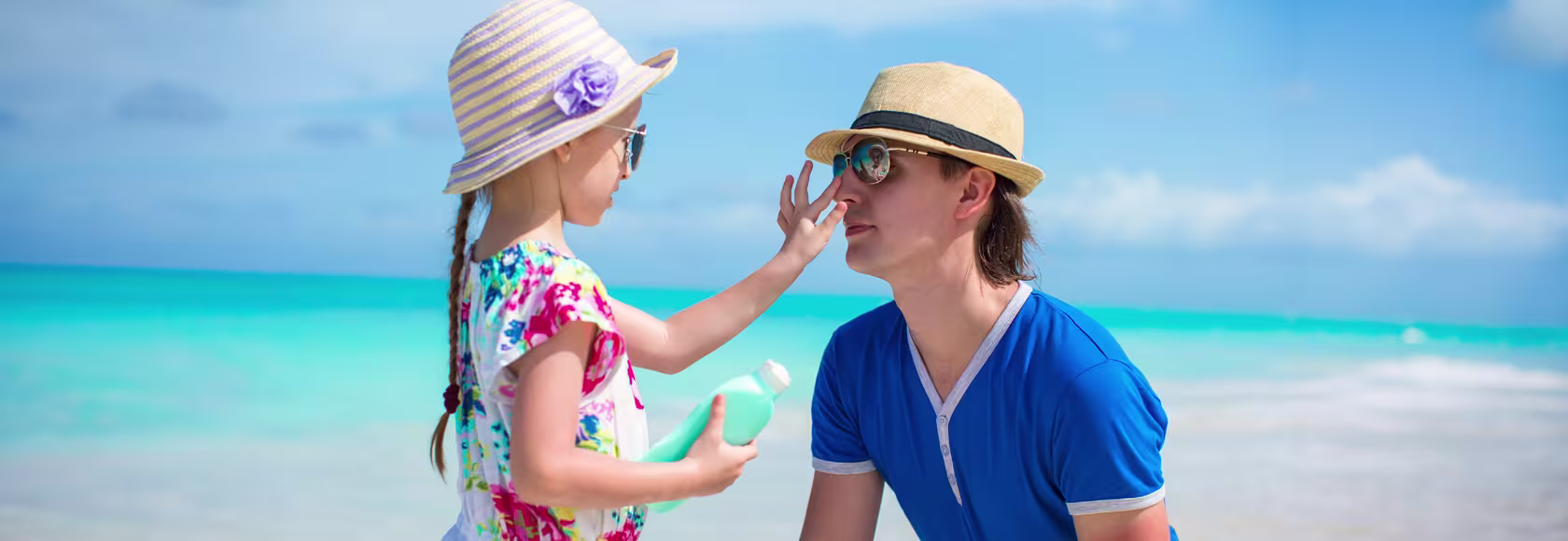 Ojciec i córka na plaży, oboje w słomkowych kapeluszach i okularach przeciwsłonecznych. Dziewczynka trzyma butelkę, drugą ręką dotyka nosa ojca.