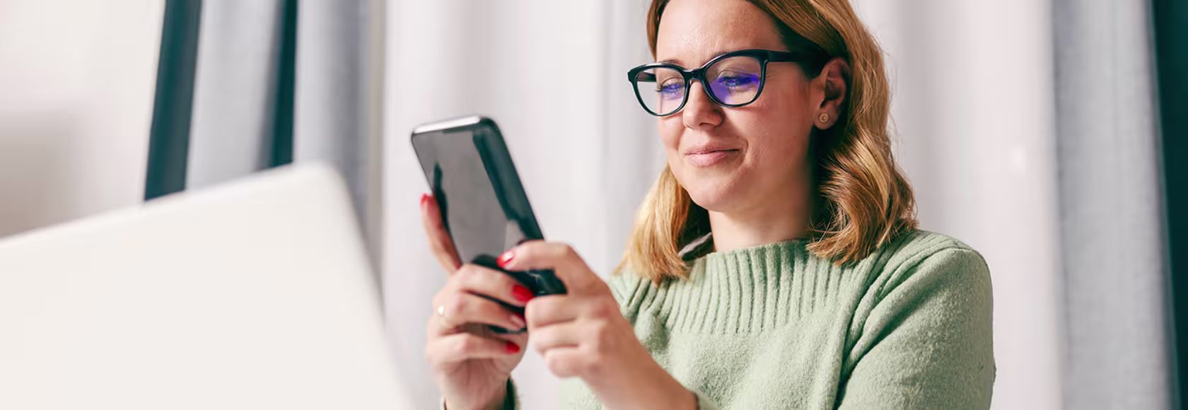 30latka z komórką, na którą patrzy, przed nią laptop