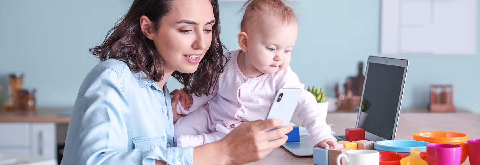Mama z niemowlakiem, trzyma komórkę, przed nią komputer