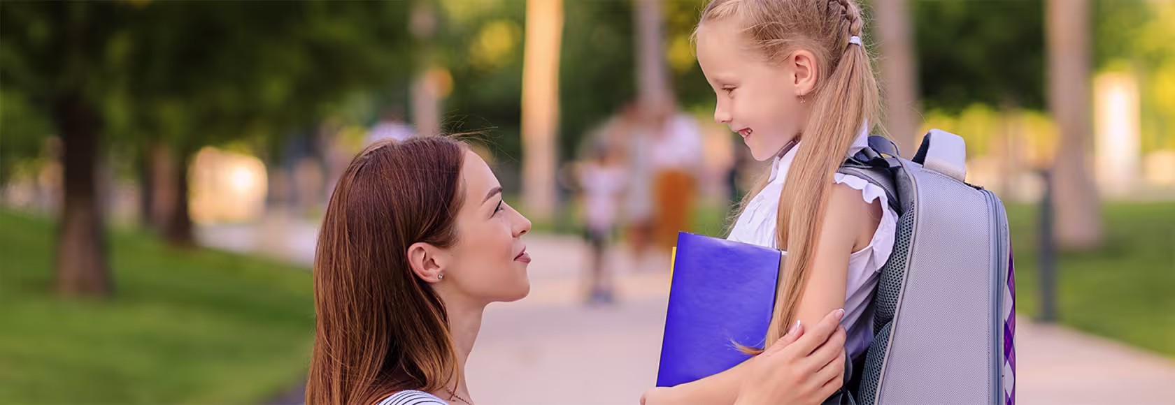 mama kuca i trzyma za ręce córkę z tornistrem, patrza na siebie z uśmiechami