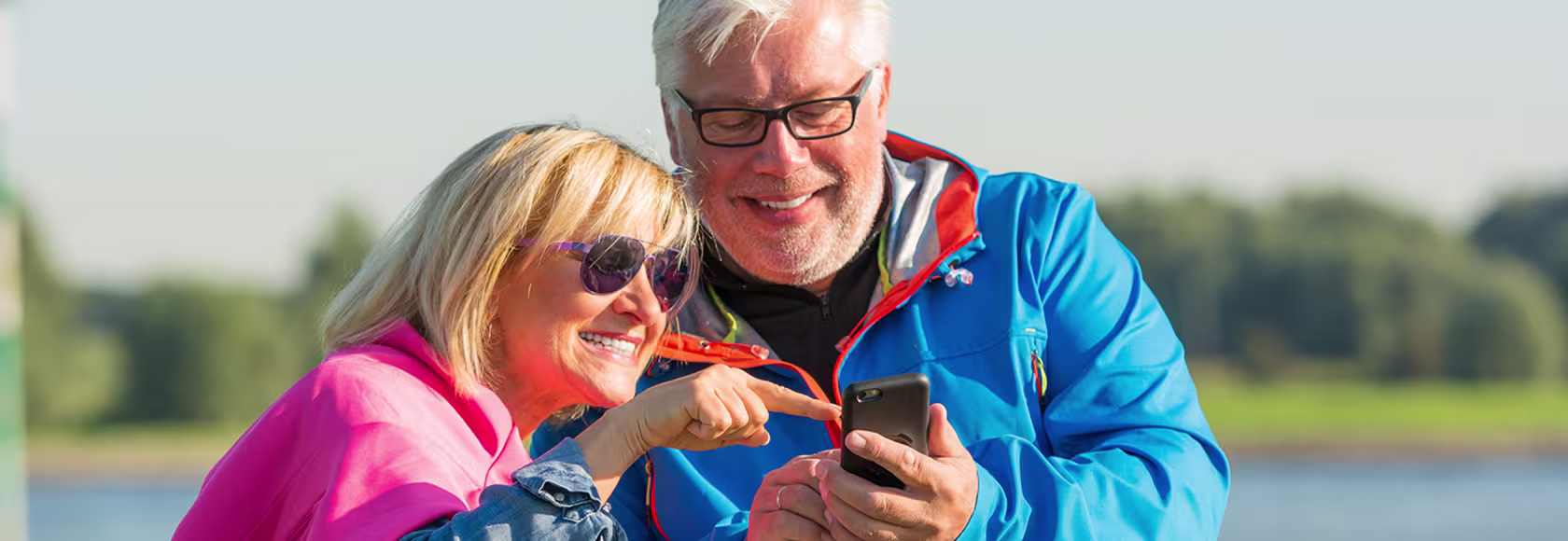 Para w średnim wieku w kurtkach nad wodą, pokazuja sobie coś na telefonie