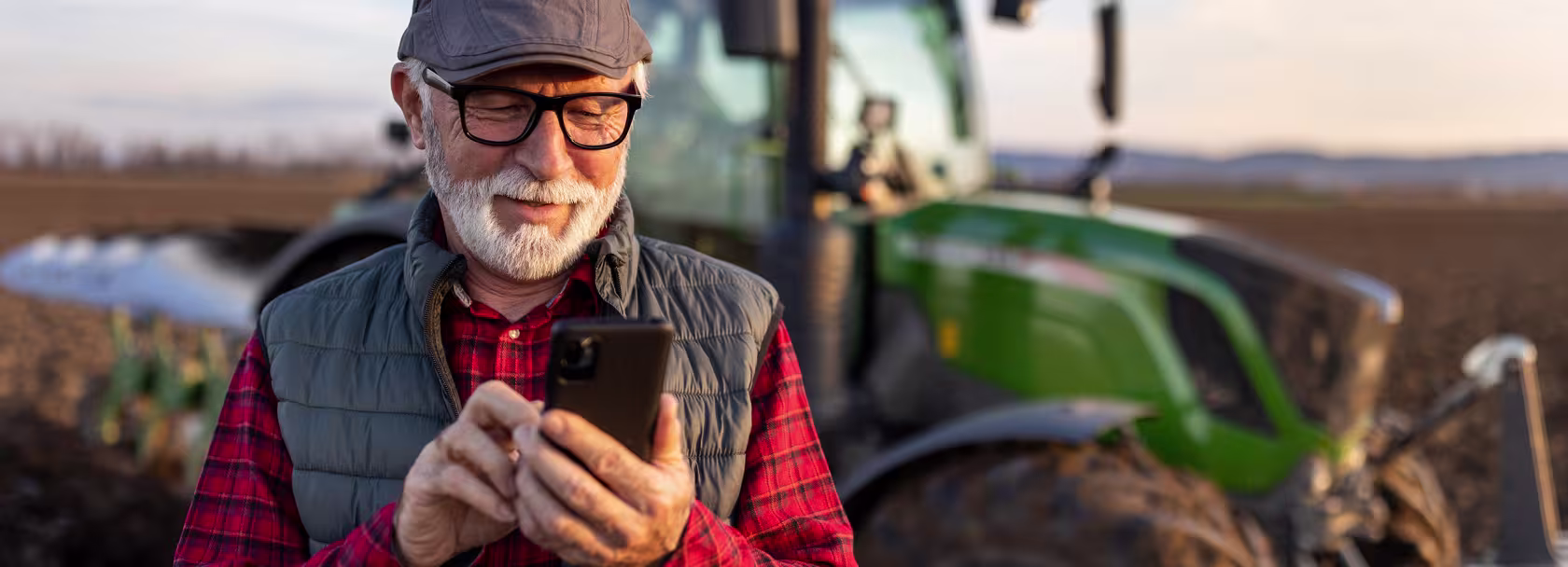 Pan z siwą brodą z komórką, w tle traktor